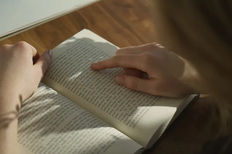 Two hands hold a book open on a wooden table; a finger follows the lines while the shadow of a plant falls across the page in afternoon light.