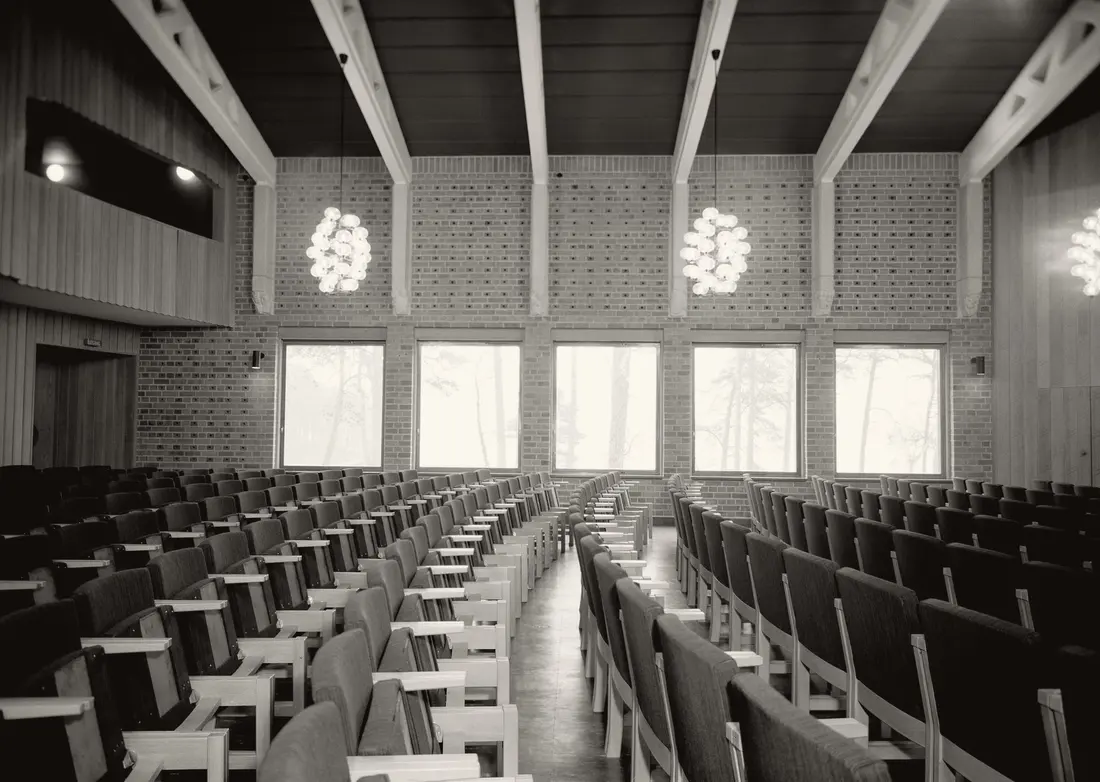 A large empty auditorium in black and white: rows of tablet-arm seats, a brick wall pierced by tall windows, pale ceiling beams and pendant lamps of clustered globes.