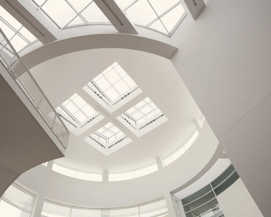 Looking up into an all-white contemporary hall: a square gridded skylight, curving balconies and soft overhead light.