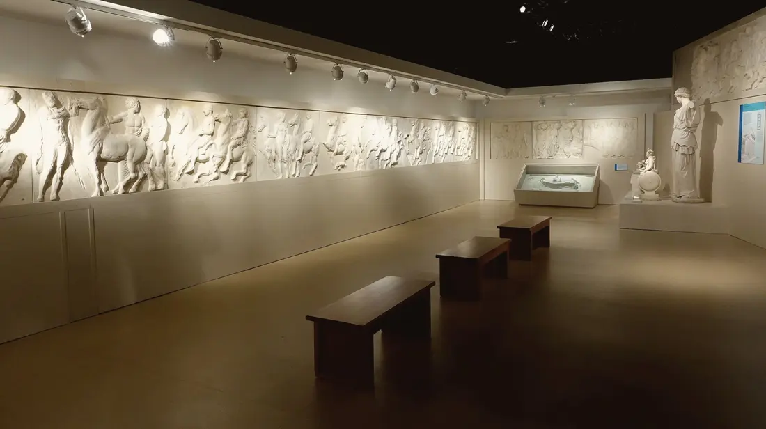 A quiet exhibition room: plaster casts of an ancient frieze lit along the wall, statues and a display case beyond, wooden benches in a row on a dark floor.