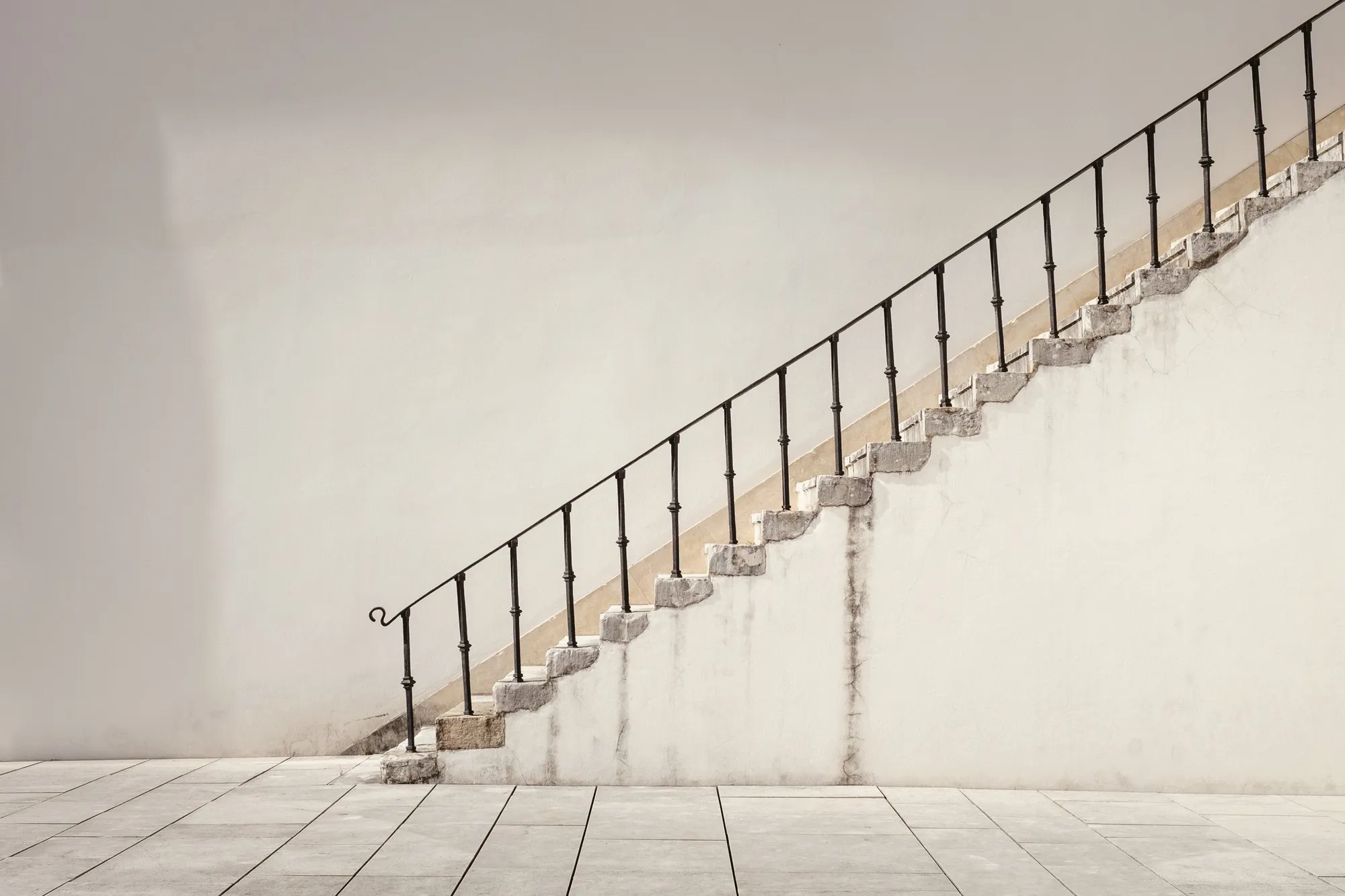 An old stone staircase climbs diagonally across a tall white wall, traced by a slender black wrought-iron handrail, above pale paving.