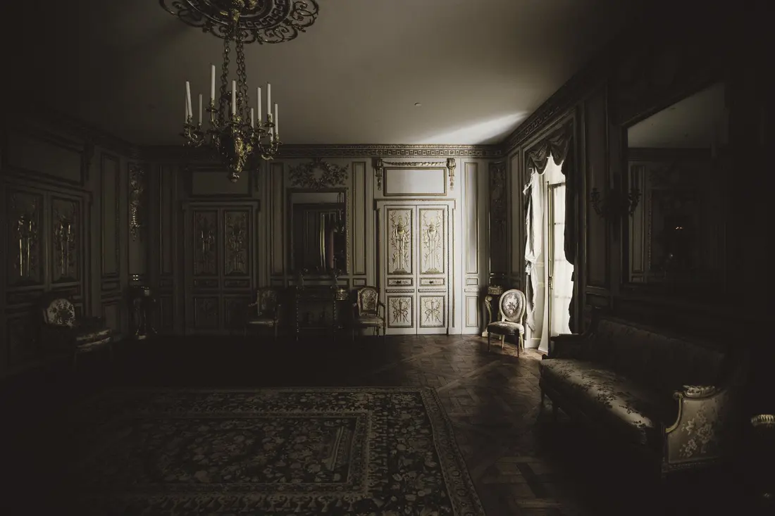 A large Louis XVI drawing room in half-light: gilded panelling, a candle chandelier, armchairs and a sofa along the walls, a Versailles parquet floor; light from a French window slides across the floor.