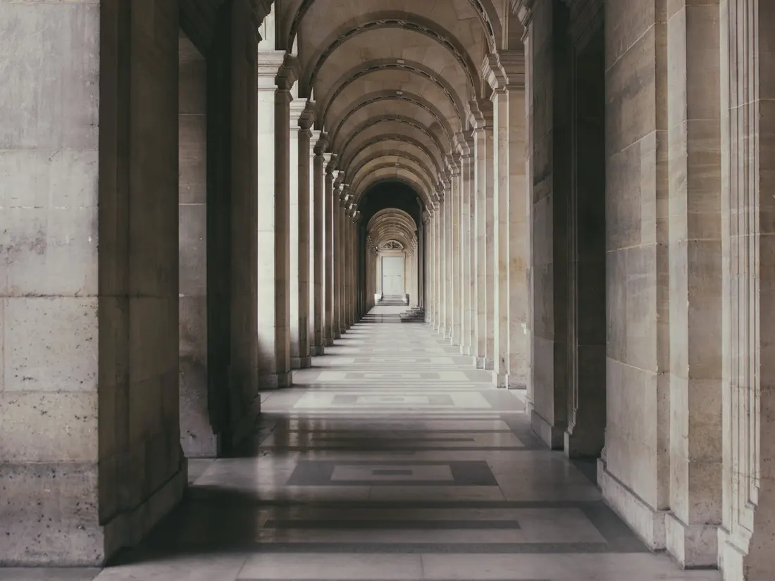A long vaulted gallery at the Louvre: a receding run of pale stone arches, marble paving and soft light to the far end.