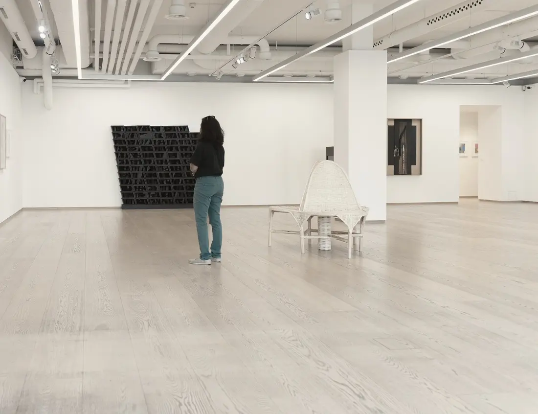 A visitor, seen from behind, pauses before a dark work in a vast white gallery with pale wooden floors; a wicker seat and a few works punctuate the space.